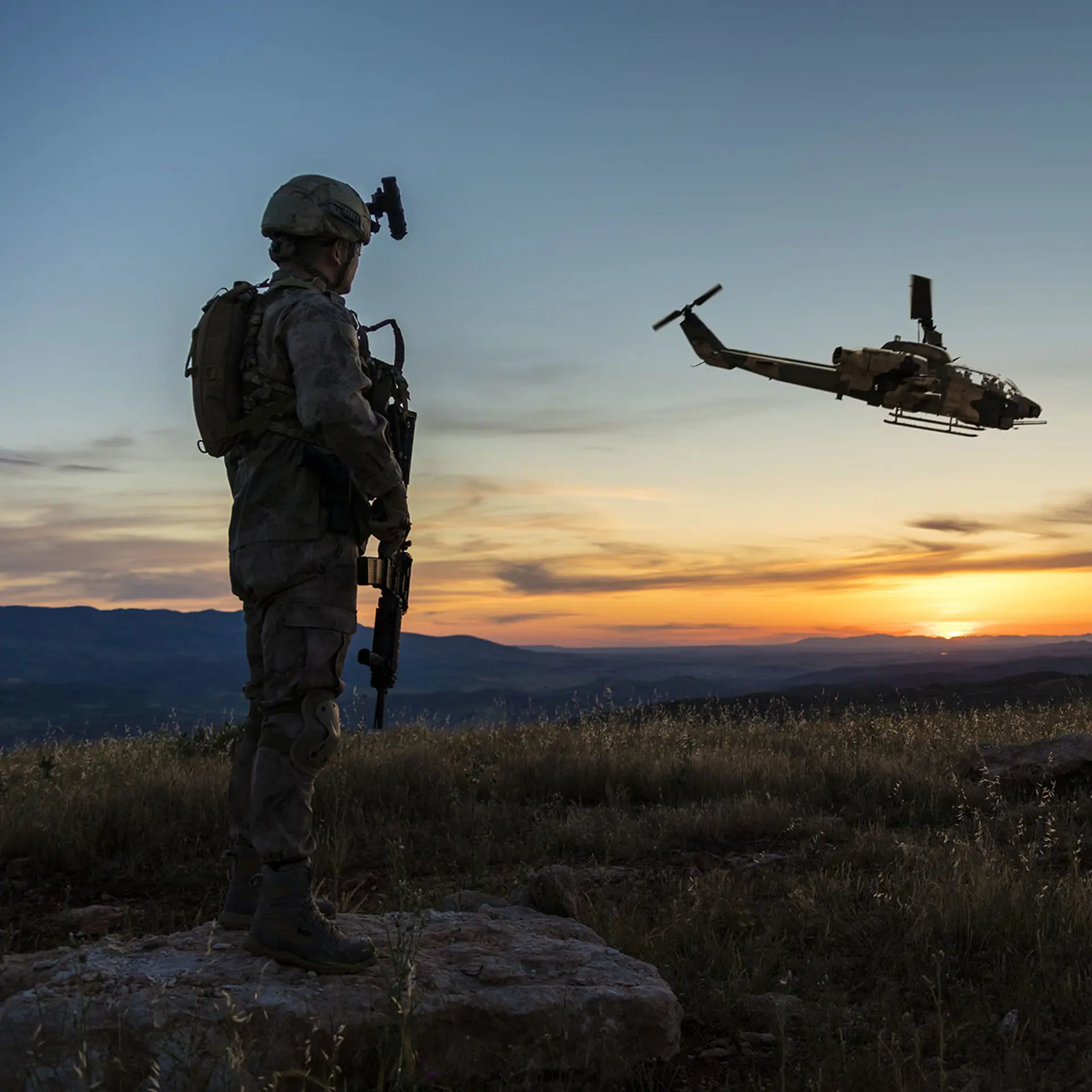 Ein Soldat in voller Kampfausrüstung steht auf einem felsigen Hügel bei Sonnenuntergang und beobachtet zwei Militärhubschrauber im Flug.