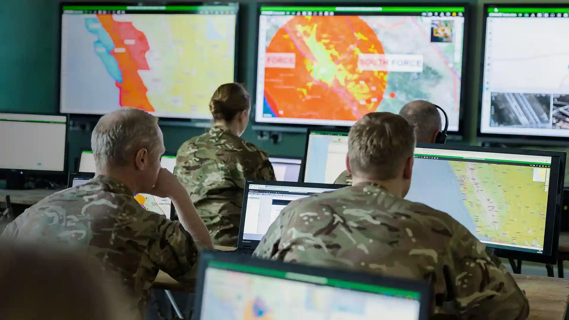 Military personnel in camouflage uniforms analyzing strategic maps and data on computer screens and large monitors in a command center, using SitaWare Headquarters.